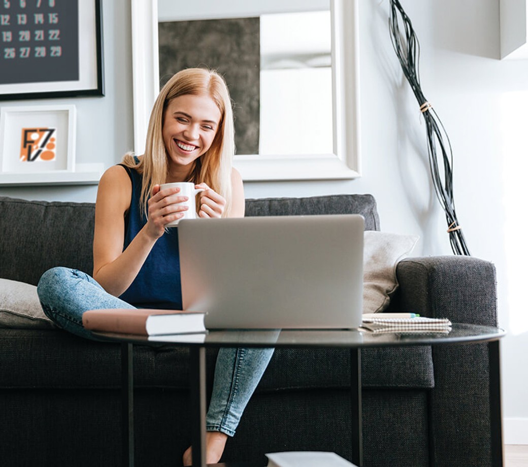 Smiling woman holding a coffee cup and looking at a laptop sitting on a sofa