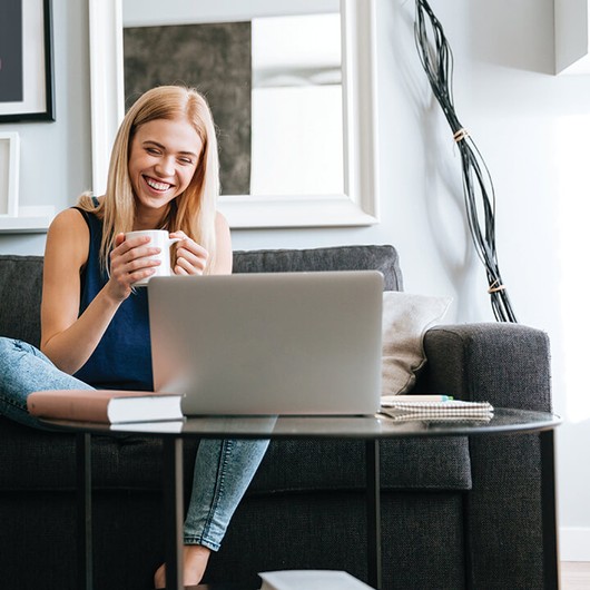 Smiling woman holding a coffee cup and looking at a laptop sitting on a sofa