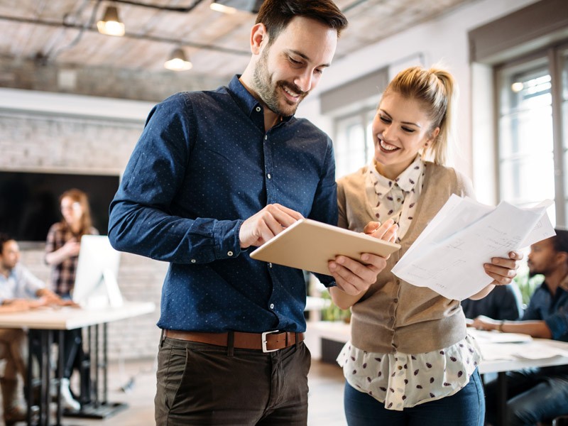 Two employees standing in the middle of an office, smiling and looking at a tablet