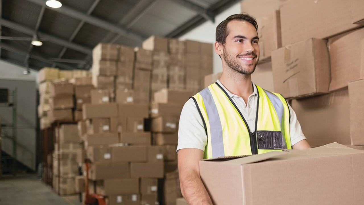 A smiling man carrying trade goods in a warehouse against a backdrop of packages