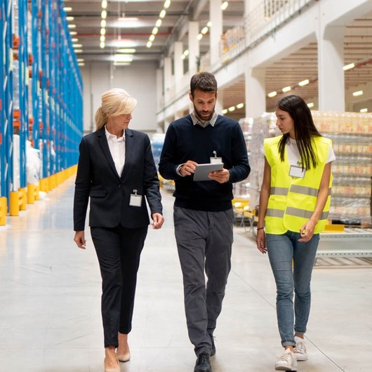 Female warehouse worker in a yellow high visibility vest walking through warehouse and assessing stock figures on a tablet with two managers