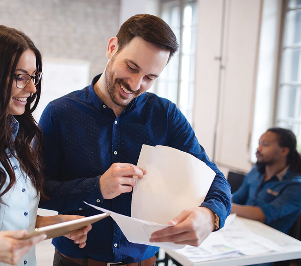 Two employees standing in the middle of an office, smiling and looking at a tablet and some documents